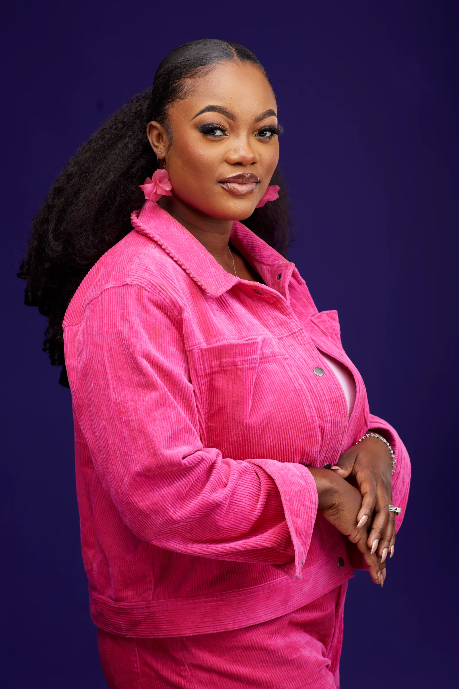 Deborah Lukalu de trois quarts, bras croisés, ensemble de velours rose sur fond violet.