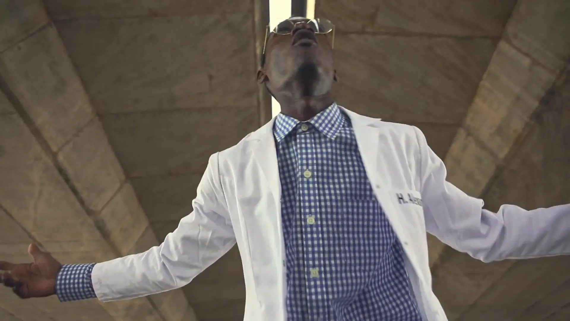 Contre-plongée : un personnage en blouse blanche ouvre les bras. Image extraite du clip.