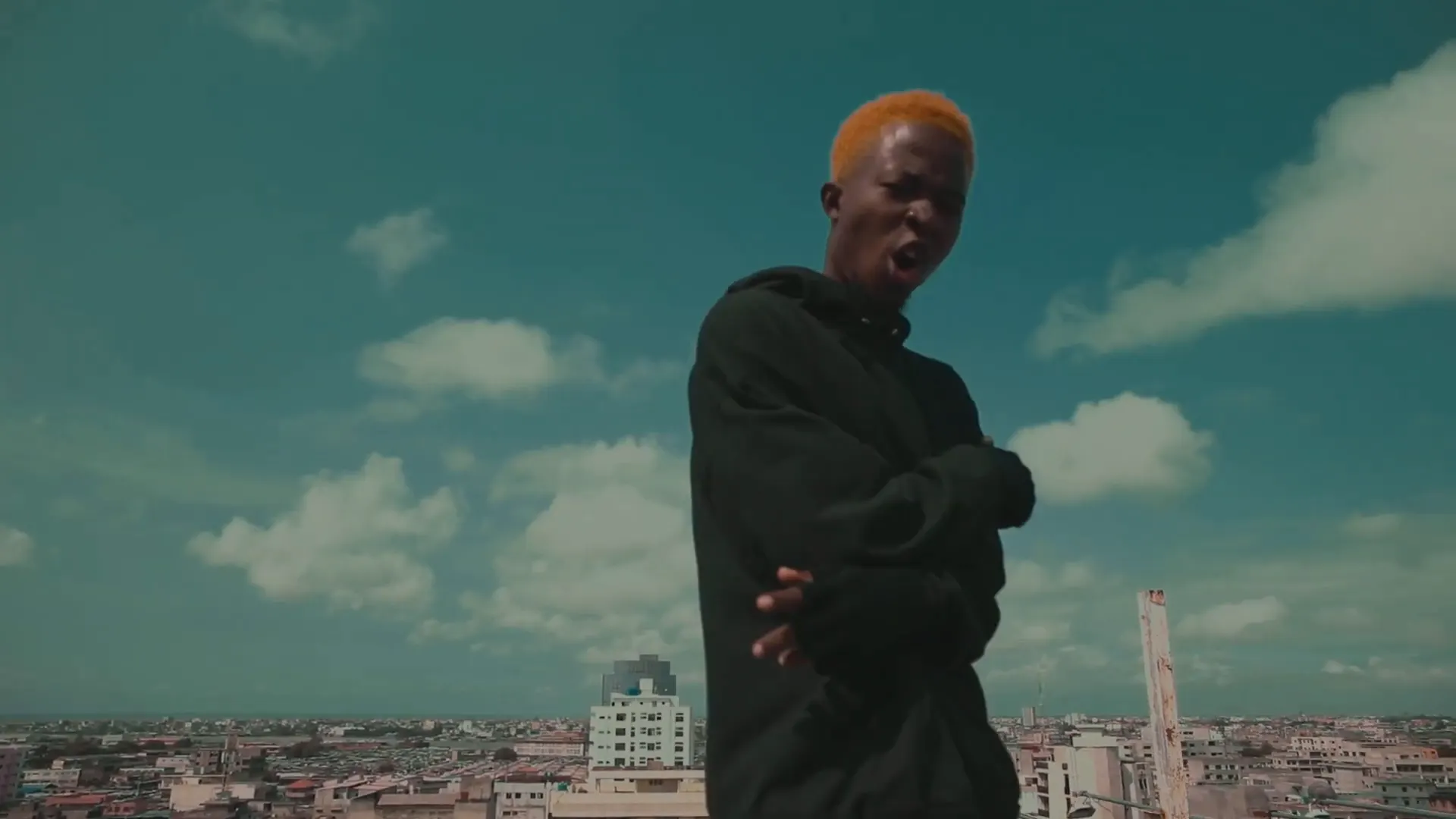 Un artiste sur un toit, ciel turquoise, la ville au loin. Image extraite du clip.