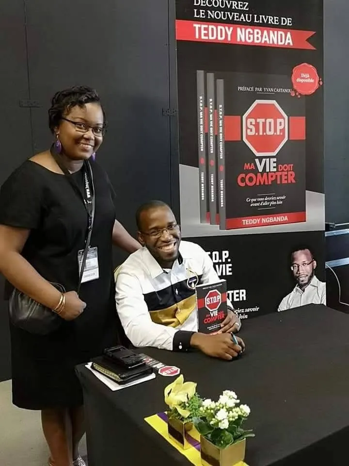 L’auteur dédicace le livre sur un stand, une lectrice à ses côtés, kakémono « le nouveau livre de Teddy Ngbanda ». Photo de terrain de la campagne.